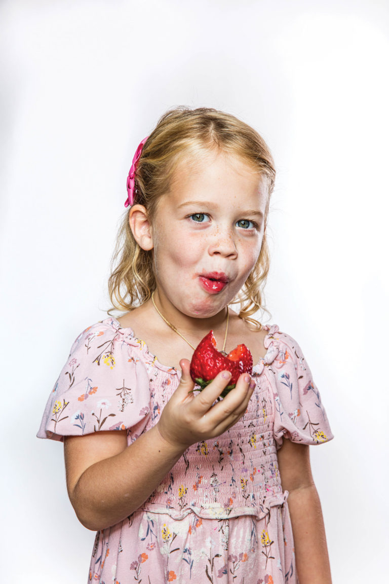A little girl eating a strawberry with a smile on her face.