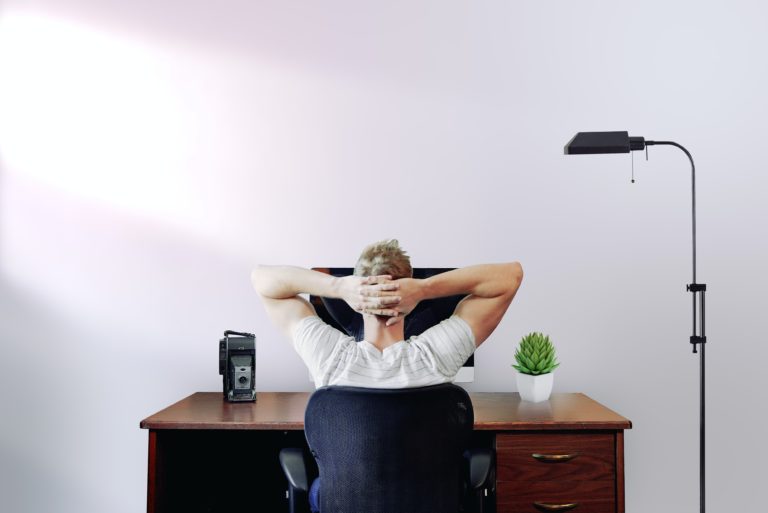 A man sitting at the computer with his hands behind his head.