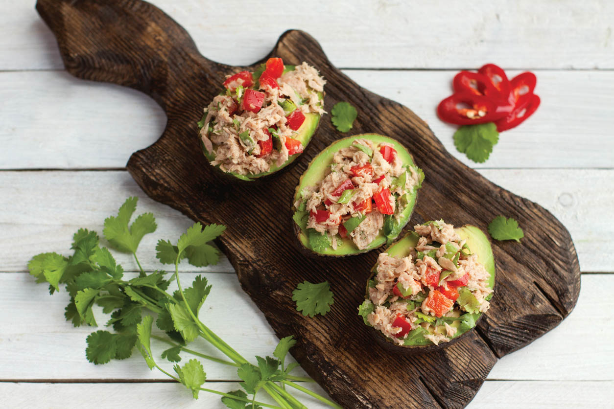 Avocado appetizers stuffed with canned tuna, bell pepper, herbs on wooden cutting board.