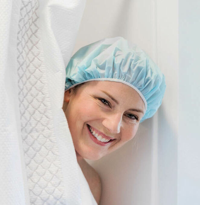 A woman smiles as she peeks out of a shower stall wearing a shower cap.