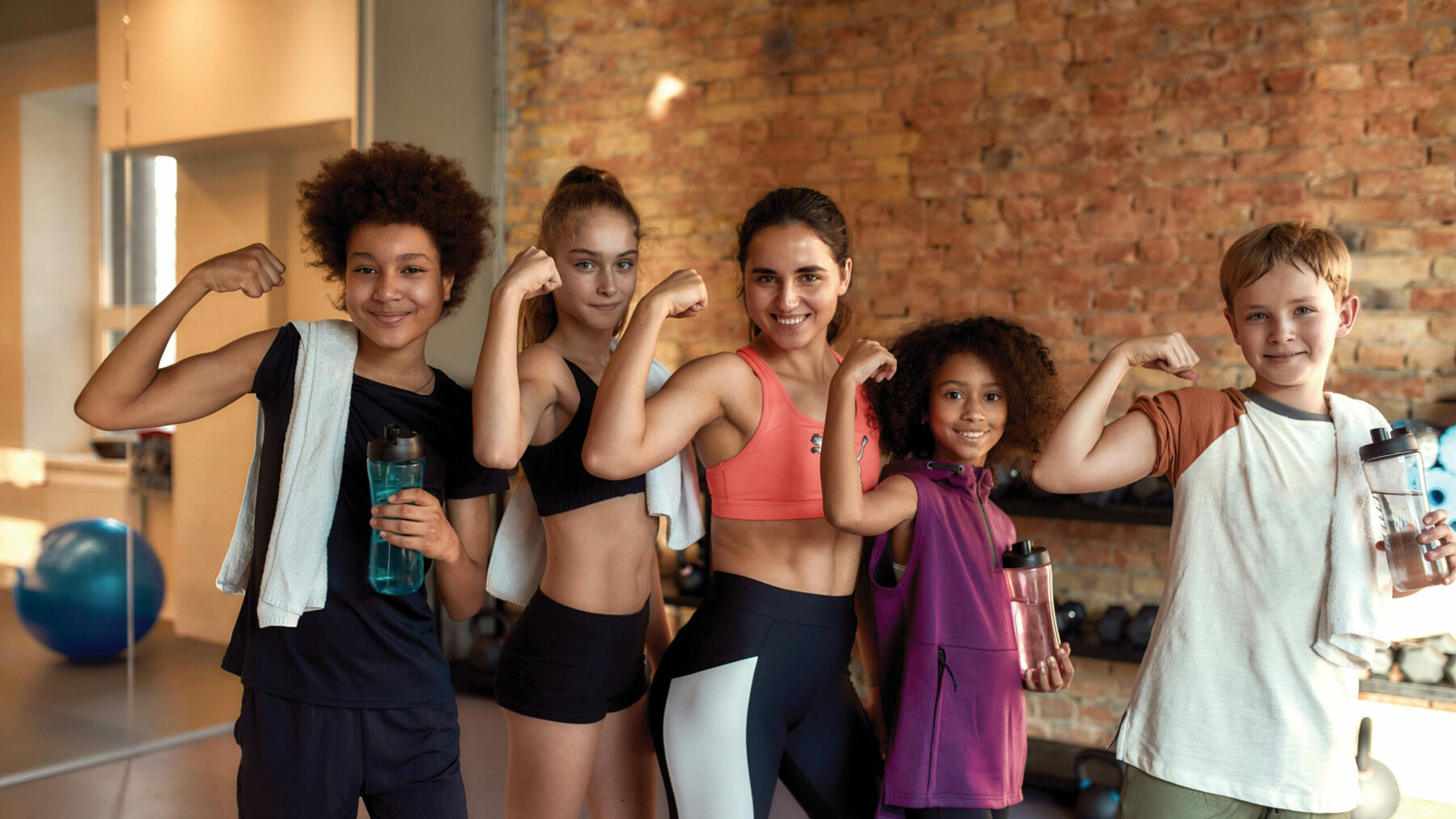 We are strong. Happy female trainer and positive children showing muscles while smiling at camera, standing in gym.